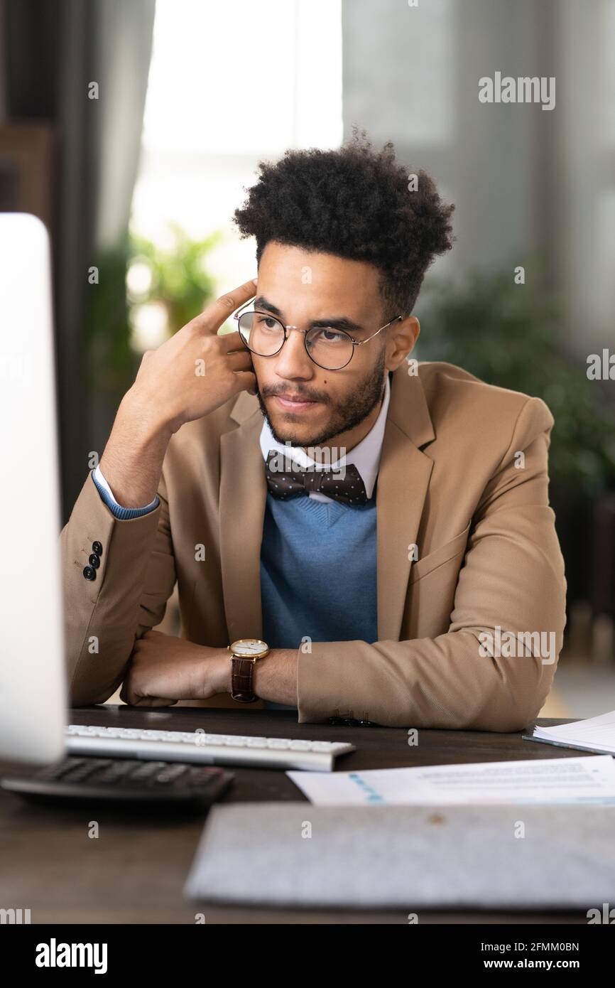 Pensive young African-American target specialist with curly hair sitting at table and leaning head on hand while analyzing research data on computer Stock Photo