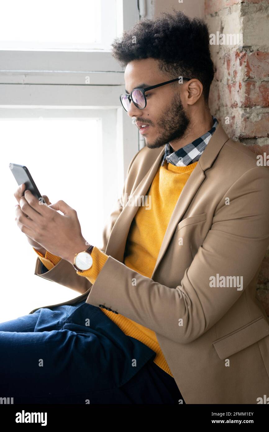 Serious young bearded man in eyeglasses sitting on window sill and texting message on phone while working online Stock Photo