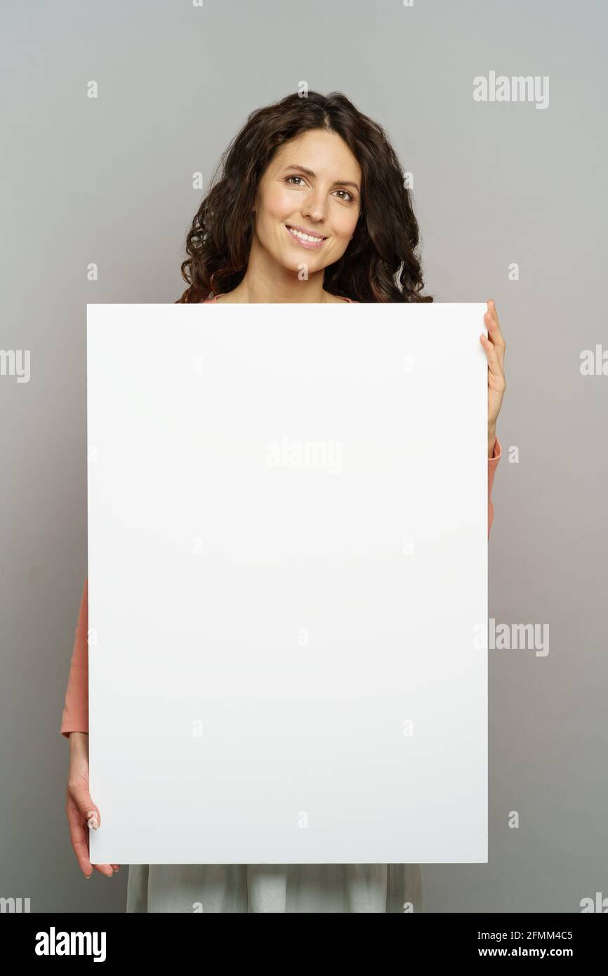 Happy millennial woman hold white blank billboard for advertising isolated at grey studio background Stock Photo