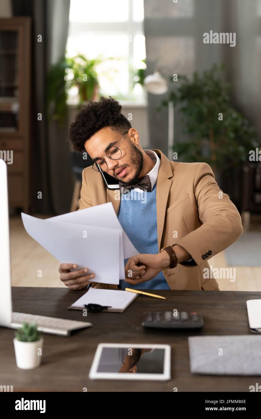Serious multi-tasking mixed race manager in bow tie sitting at table with calculator and checking time while talking by phone and examining papers. Stock Photo