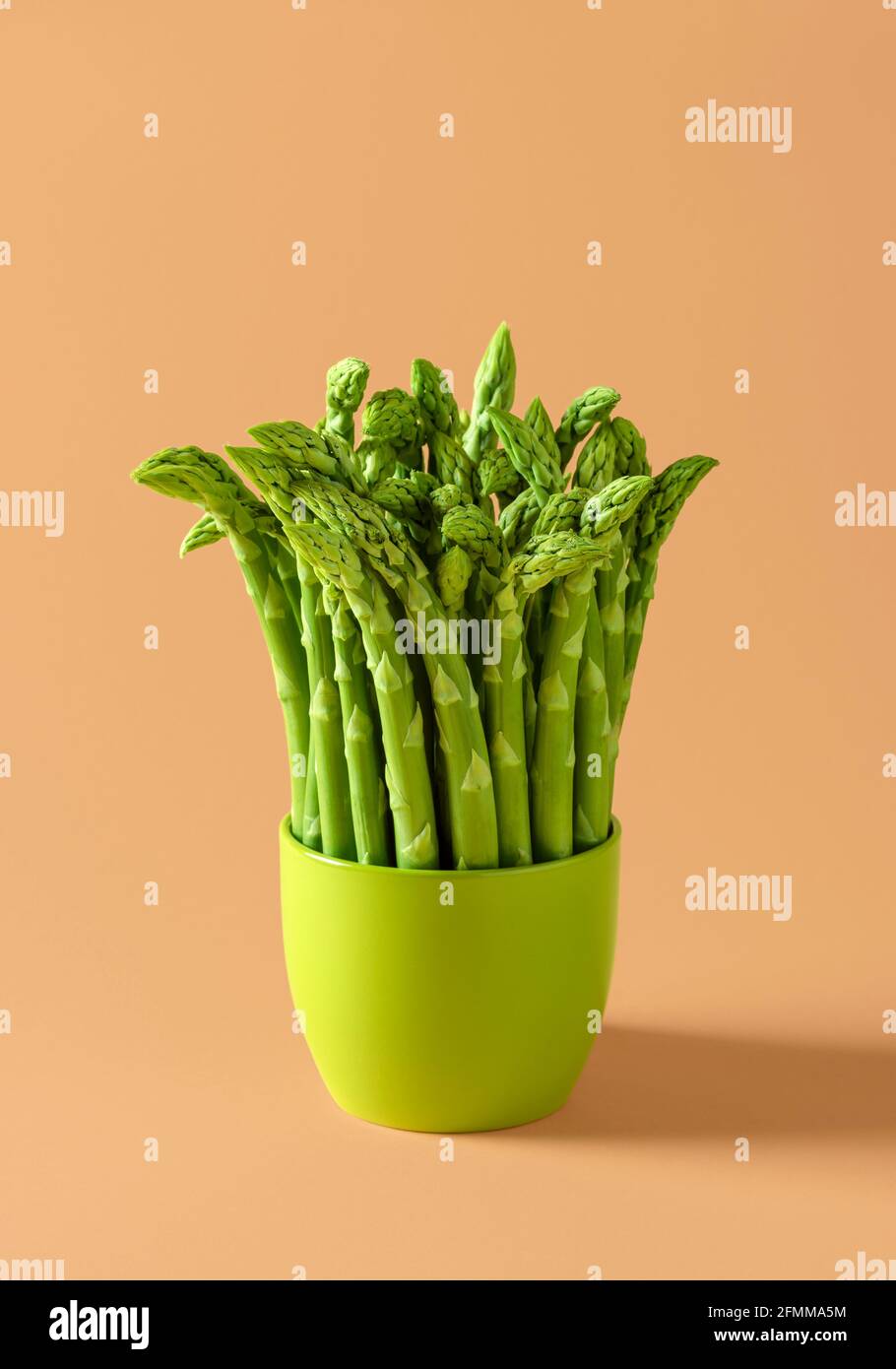 Bunch of raw asparagus in a green ceramic pot on a beige background. Fresh green asparagus on a colored background. Stock Photo