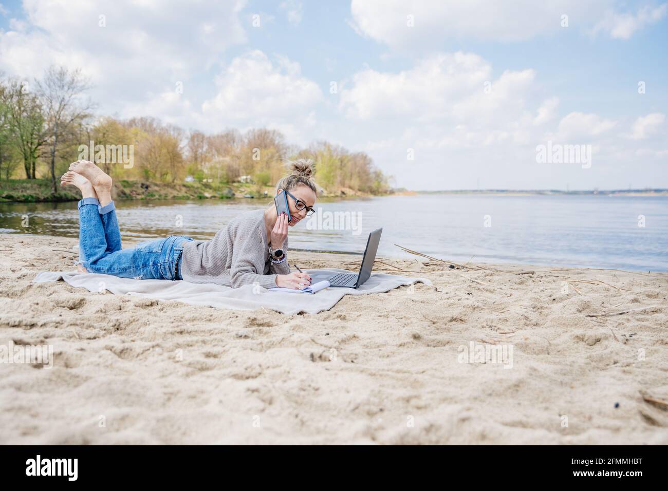 Woman works outdoors on laptop. She's lying on the beach on a blanket and talking on the phone with klient. Stock Photo