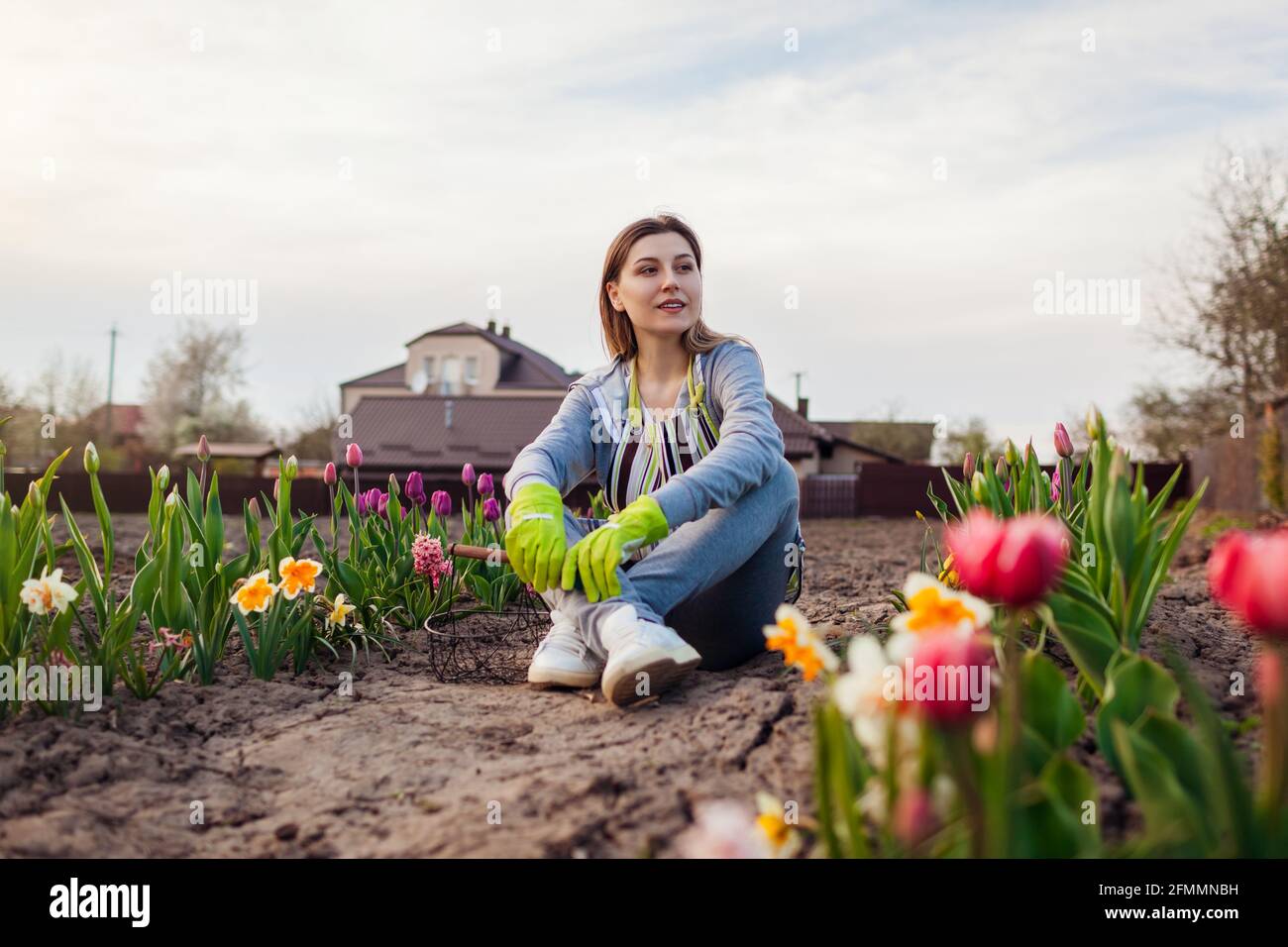 Young gardener relaxing among fresh tulips, daffodils, hyacinths in spring garden. Happy woman enjoys colorful flowers sitting on the ground Stock Photo