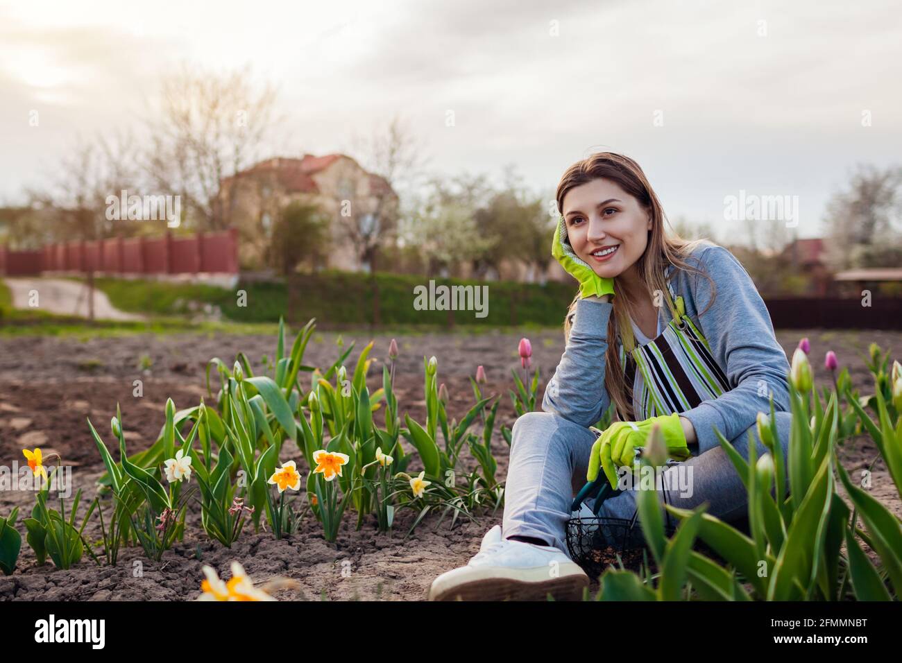 Smiling gardener relaxing among fresh tulips, daffodils, hyacinths in spring garden. Happy woman admires colorful flowers sitting on the ground Stock Photo