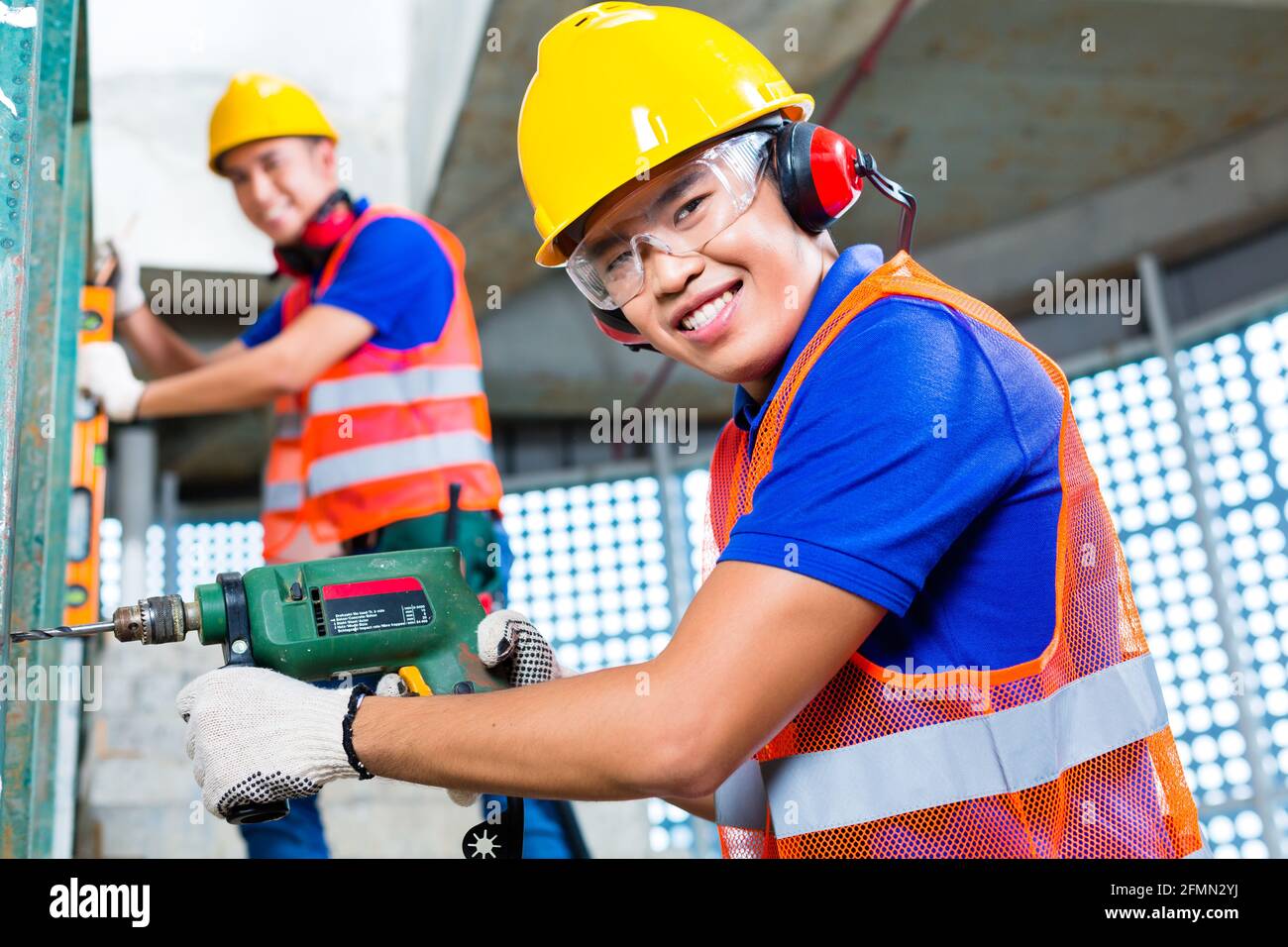 Asian Indonesian construction site workers  drilling with a machine or drill, bubble level, ear protection, gloves and hardhat or helmet in a wall of Stock Photo
