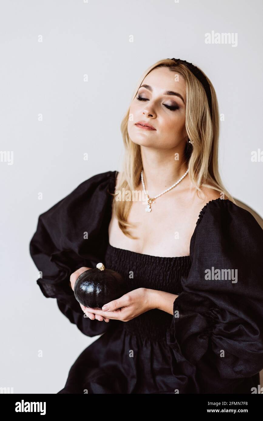 Beautiful blonde woman in a black dress with lush sleeves and a pumpkin in her hands. Halloween trendy outfit. Soft selective focus, artistic noise. Stock Photo