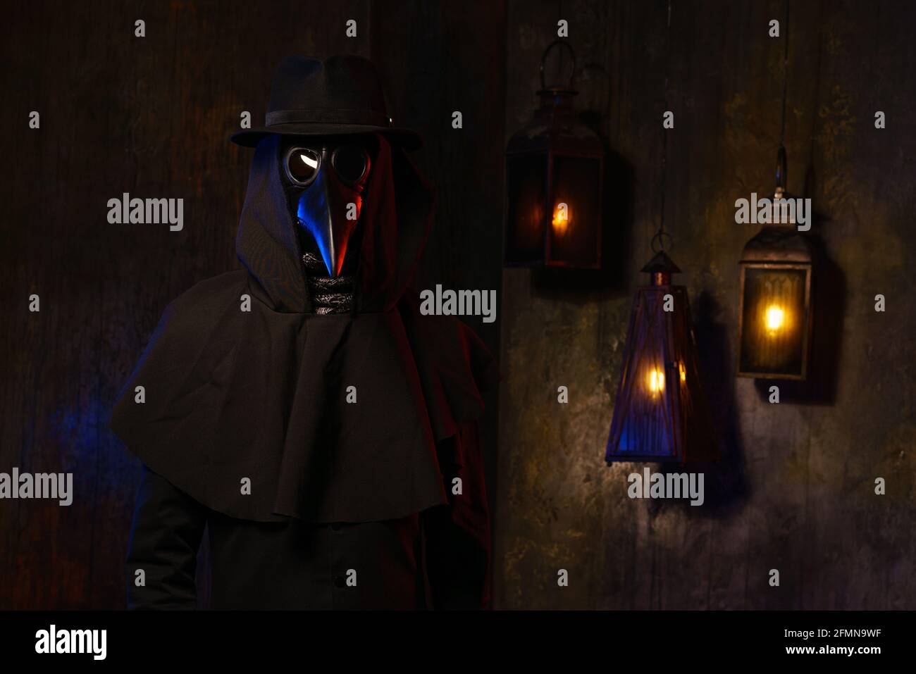 Portrait of a plague doctor in a black leather mask, hat and hood, isolated against a dark wall with lanterns. Stylization of a historical costume, a creepy image, the concept of an epidemic. Stock Photo