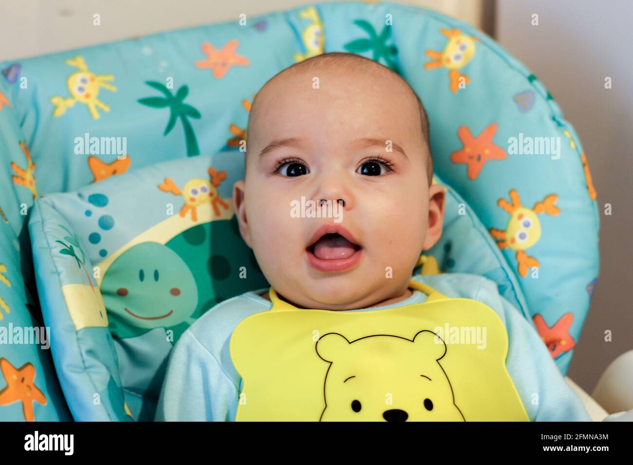 Baby in an apron sits in highchair and waits to be fed. Serious baby looking to camera Stock Photo