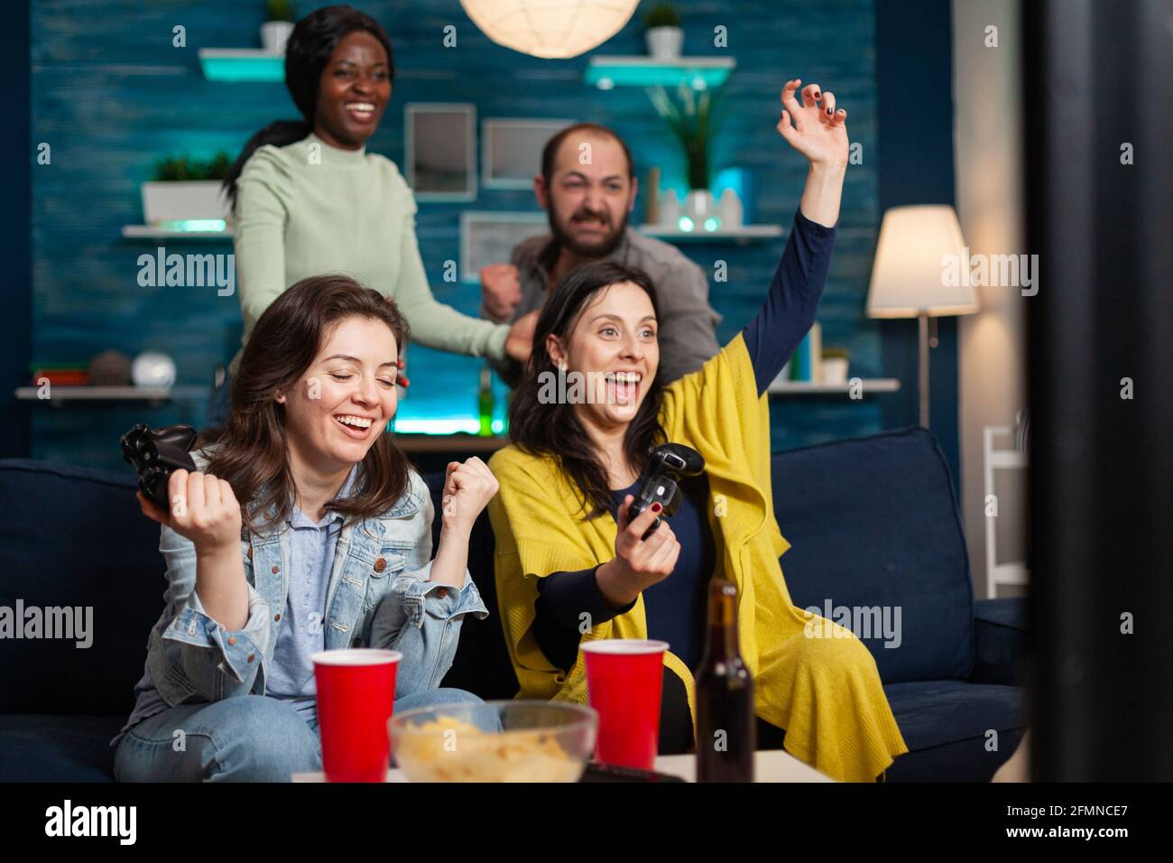 Cheerful women celebrating victory while playing video games with friends using wireless controller. Group of mixed race friends playing games while sitting on sofa in living room late at night. Stock Photo