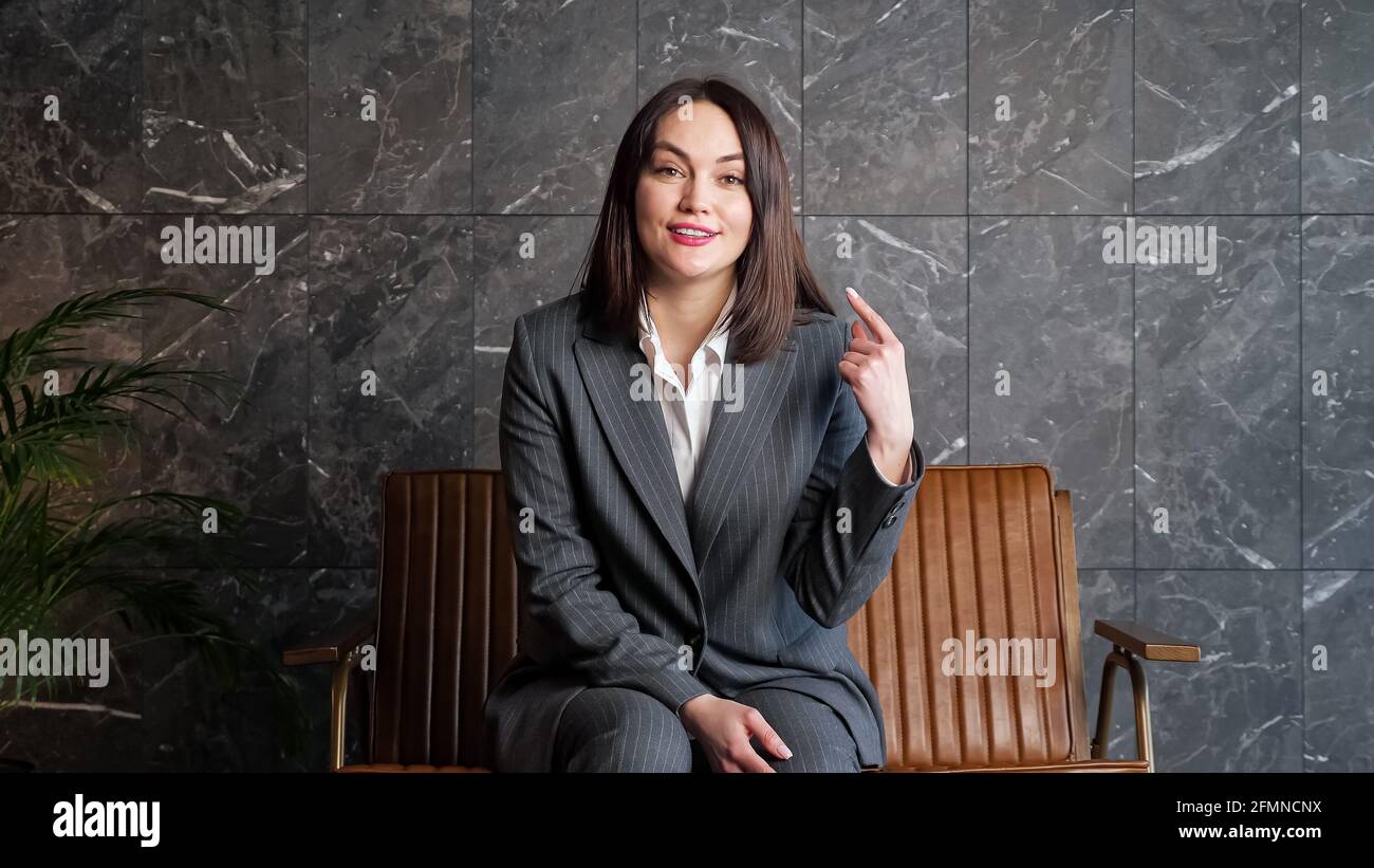 Smiling brunette in grey classic suit talks and gesticulates sitting on  brown bench near large pot plant at job interview in new office Stock Photo  - Alamy