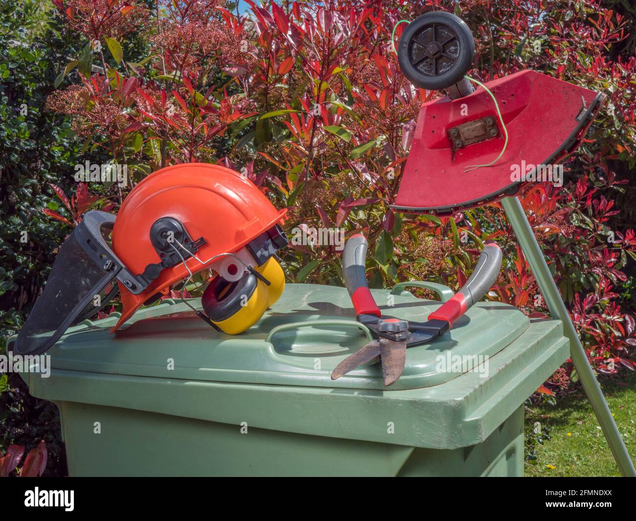 Closeup of gardening equipment – hard hat, shears and a strimmer - resting on a green wheely bin, with colourful bushes in the background. Stock Photo