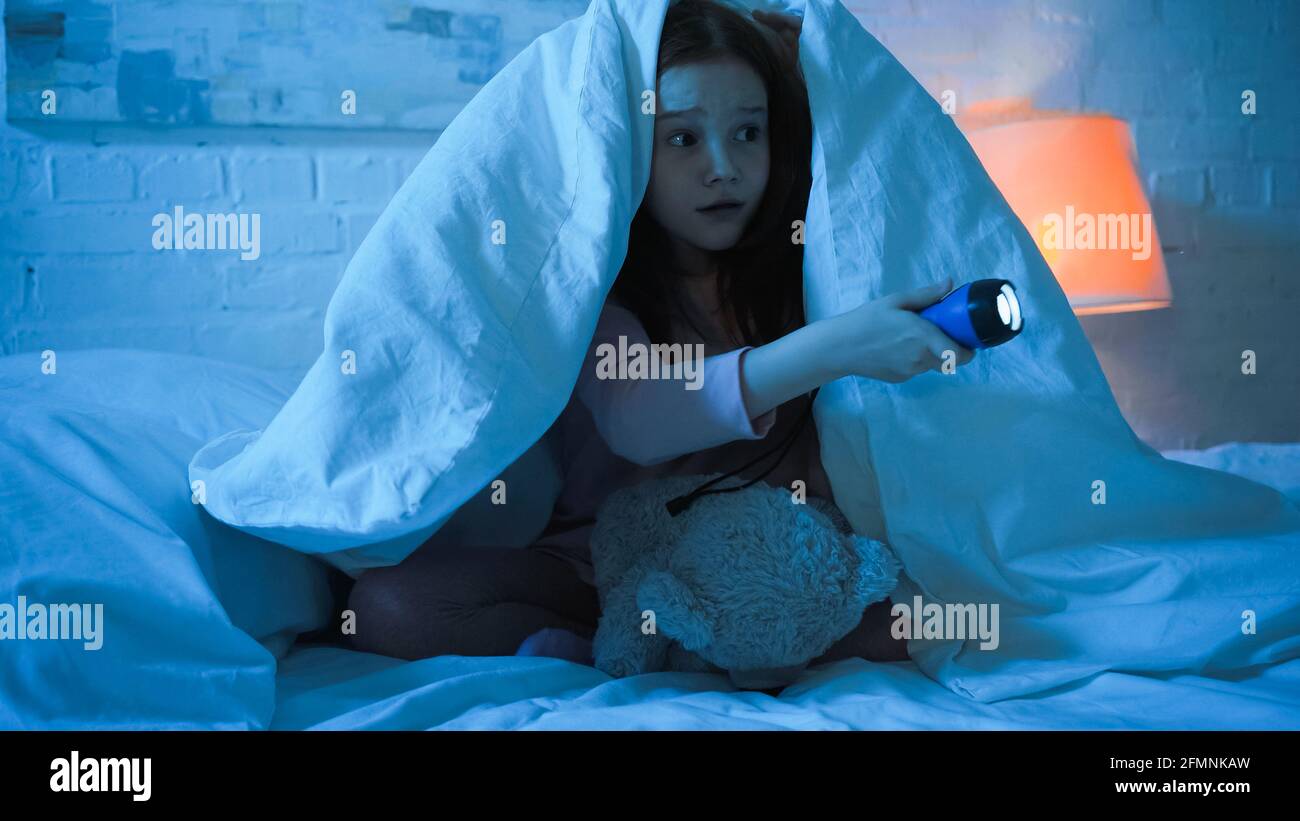 Scared kid with teddy bear holding flashlight on bed in night Stock Photo