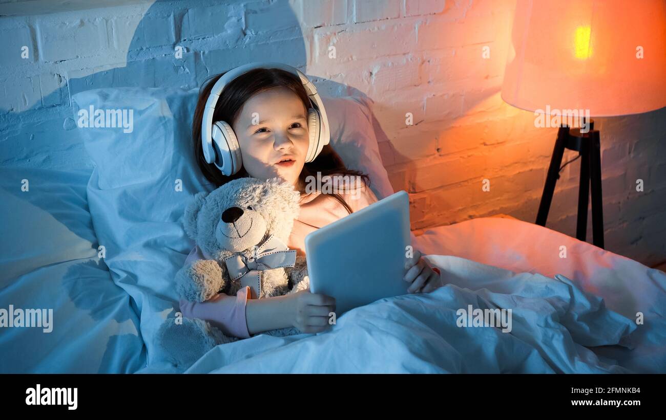 Child in headphones holding digital tablet and hugging teddy bear on bed Stock Photo