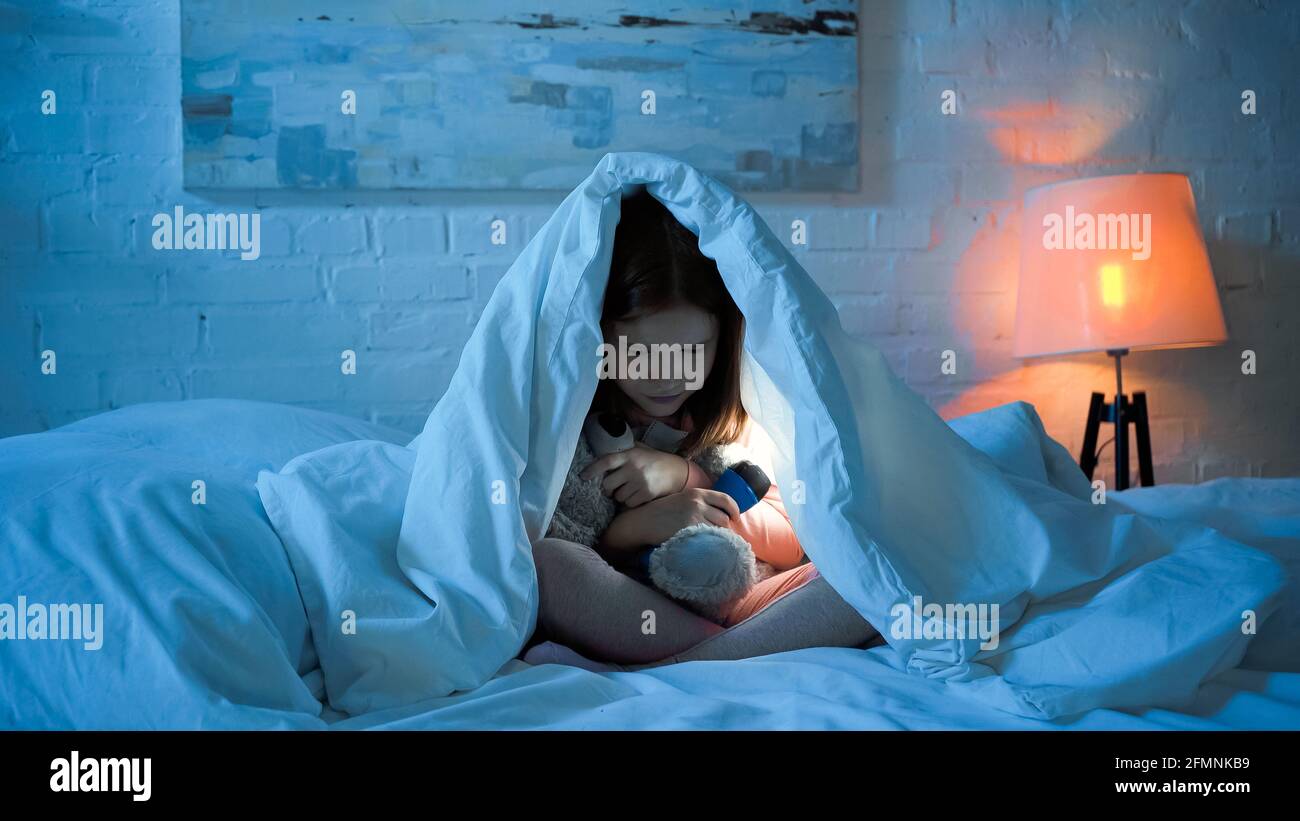 Scared girl holding flashlight and hugging soft toy on bed in night Stock Photo