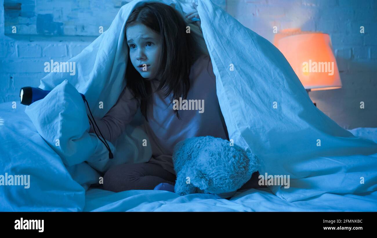 Scared child holding flashlight near teddy bear under blanket on bed Stock Photo