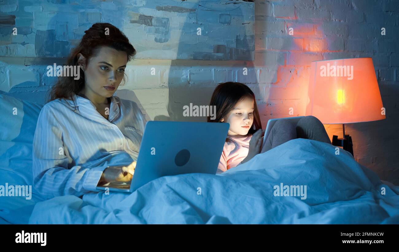 Freelancer using laptop near daughter with digital tablet on bed Stock Photo