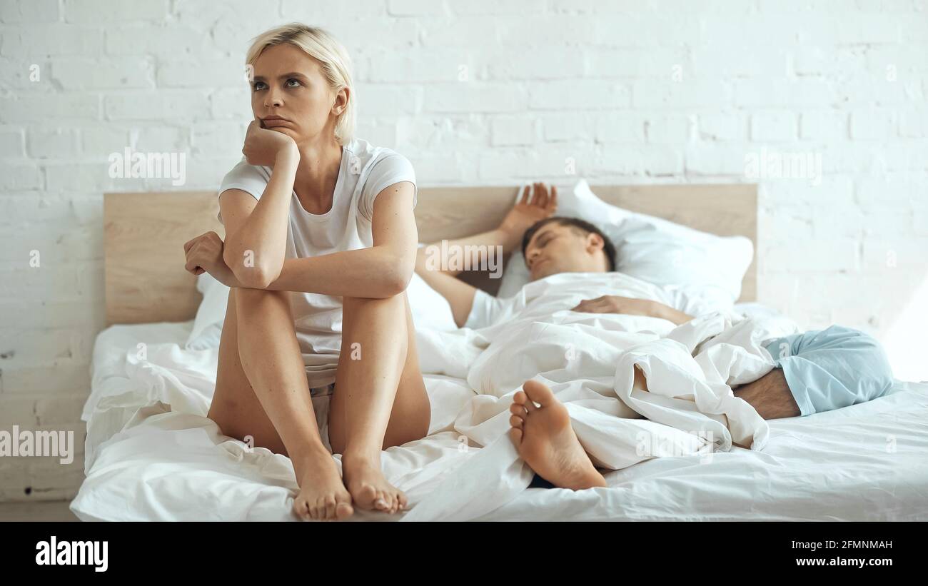 disappointed and barefoot woman sitting on bed while boyfriend sleeping in bedroom Stock Photo