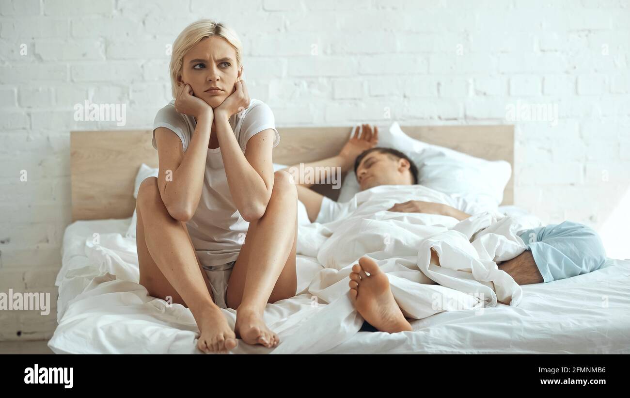 dissatisfied and barefoot woman sitting on bed while boyfriend sleeping in bedroom Stock Photo