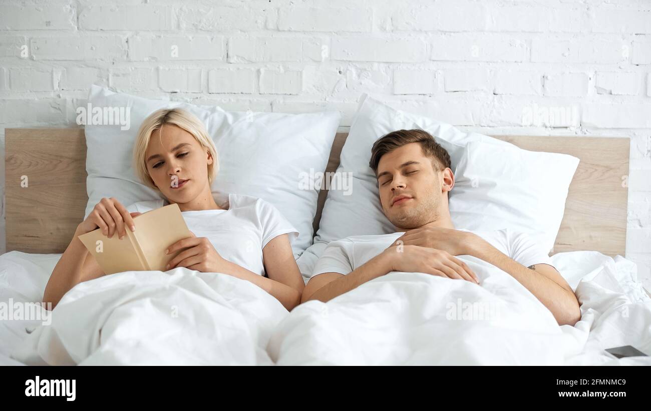 young blonde woman reading book near sleeping boyfriend in bedroom Stock Photo