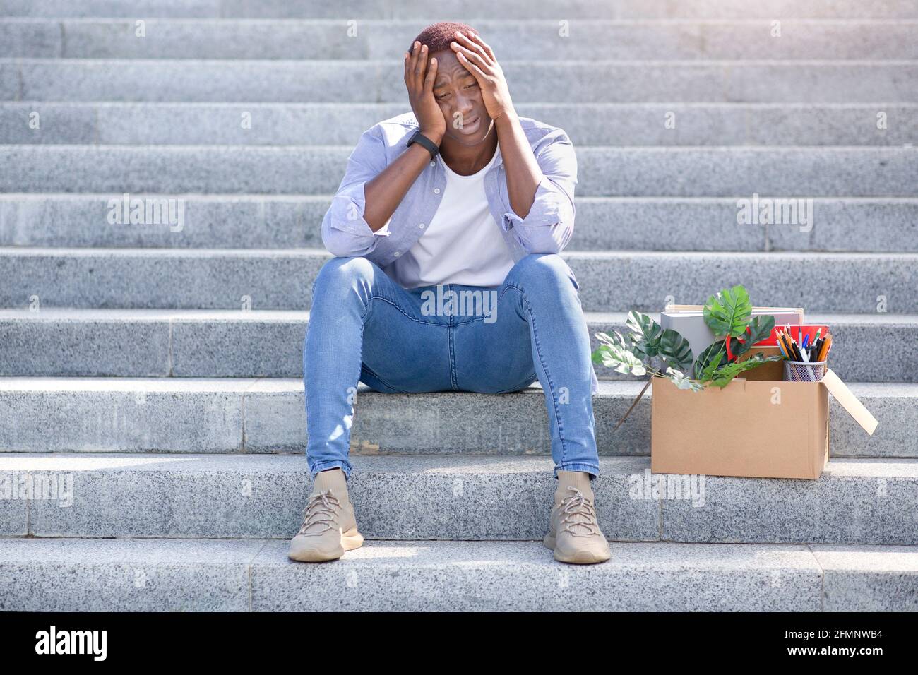 Economic crisis. Desperate African American man with personal belongings sitting on stairs outdoors after being fired Stock Photo
