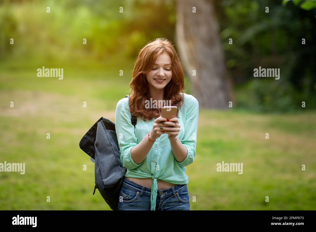 Cheerful redhead student girl using smartphone outdoors, texting with friends Stock Photo