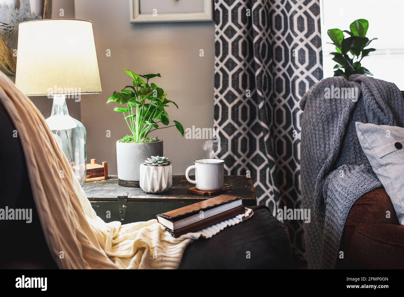 Cozy setting of a hot, steaming cup of coffee sitting on a vintage chest with houseplants and lamp in a farmhouse style living room. Stock Photo