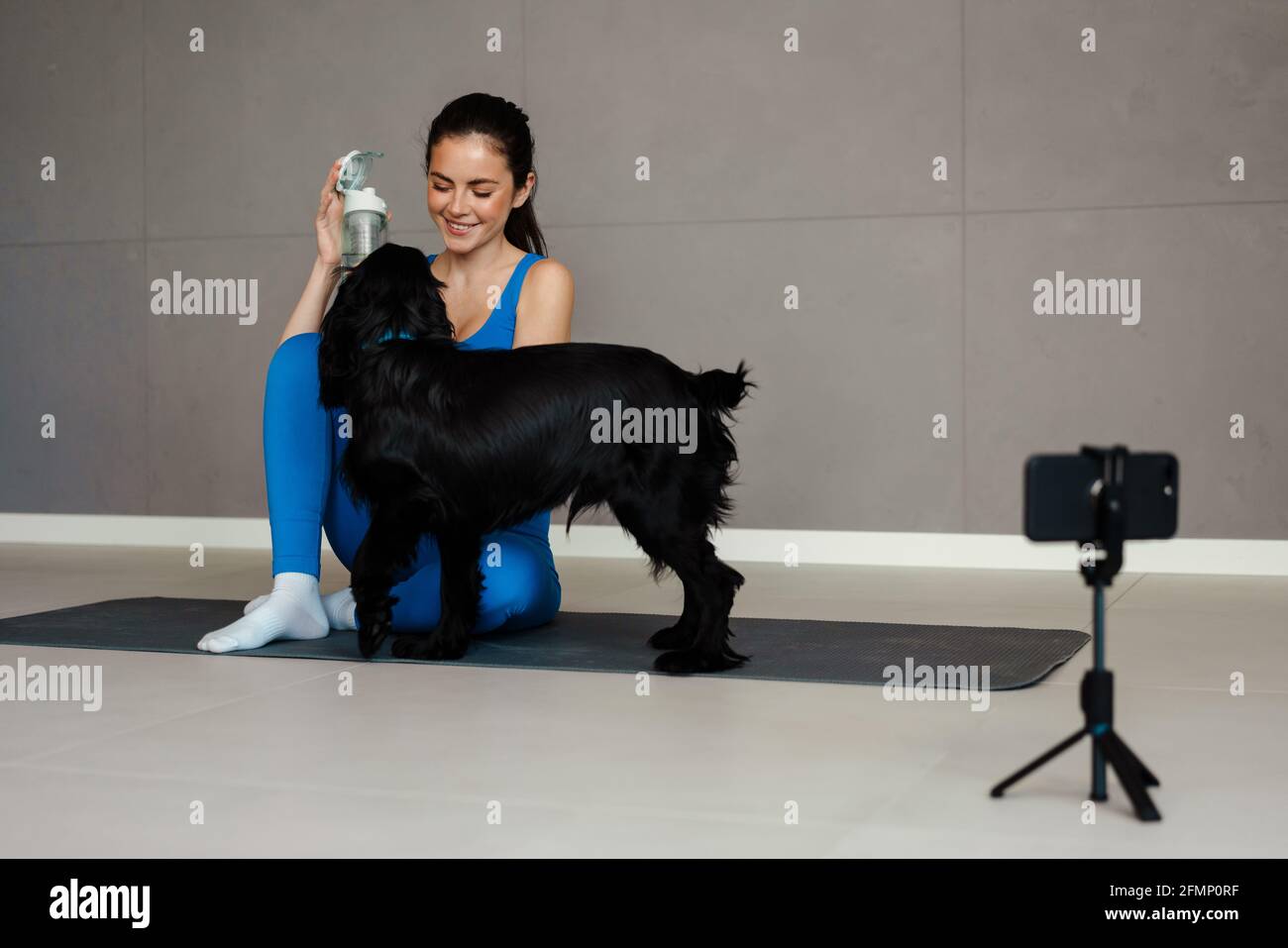 Happy young brunette woman blogger exercising with her dog on a fitness mat in front of camera indoors Stock Photo