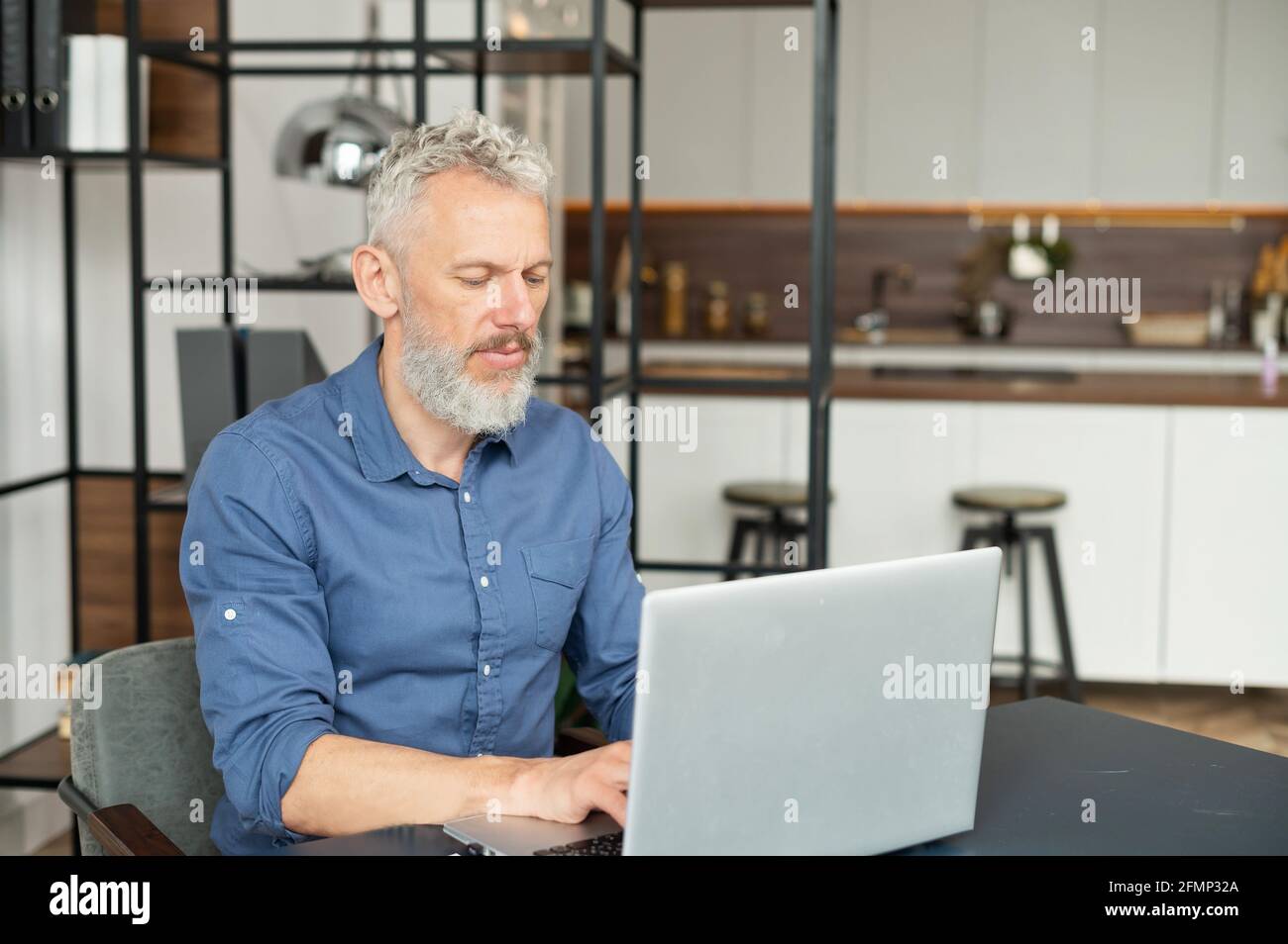 Focused mature man typing on the keyboard, senior male using laptop for remote work, messaging, working on the new startup sitting at the desk in modern apartment Stock Photo