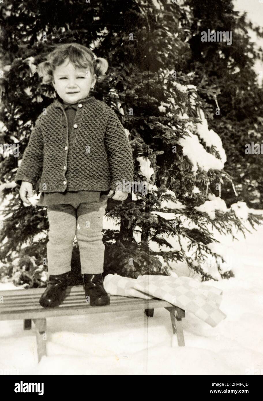 2 years old girl standing on simple wooden sleigh next to a tree in snowy winter c.1970, Bulgaria Stock Photo