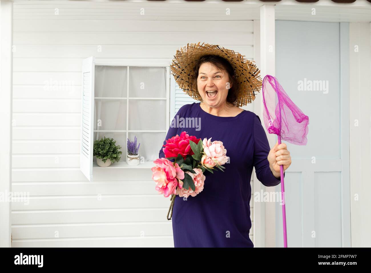 Joyful woman aged 55 in her backyard in hat with butterfly net in her hands. Concept of relaxation and unity with nature. gardening worker Stock Photo