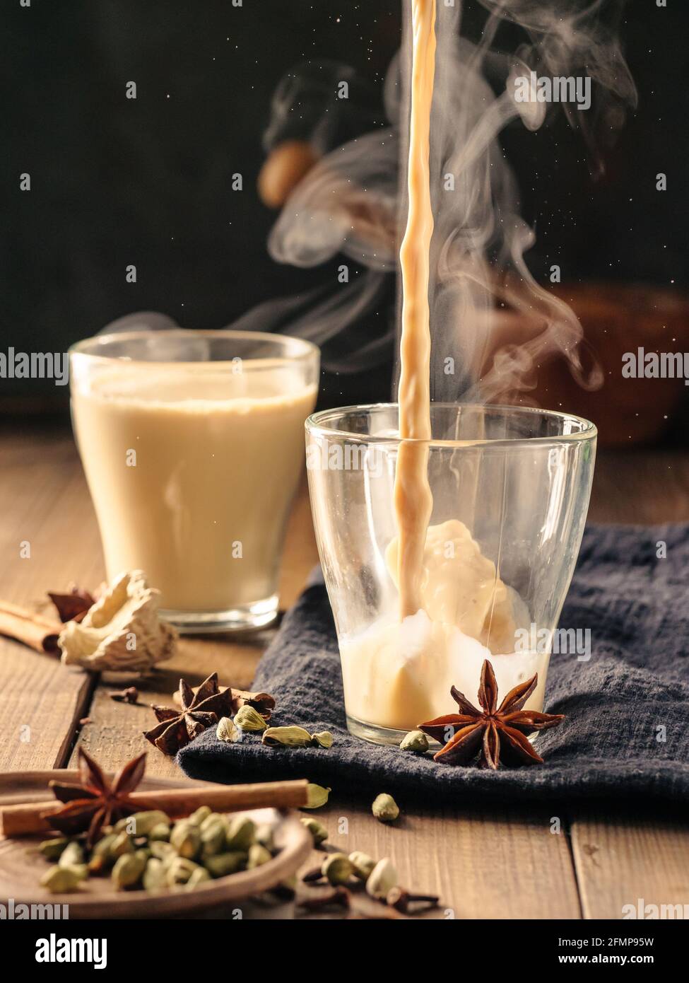 Masala tea is poured into a glass mug. Spices are laid out on the table. Stock Photo