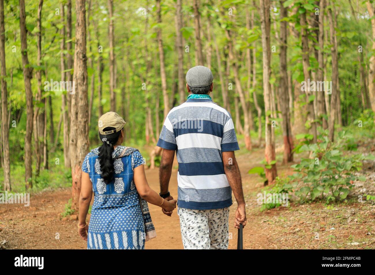 Backside view of a middle aged couple (40-45 yrs) walking hand in hand in a green dense forest wearing caps Stock Photo