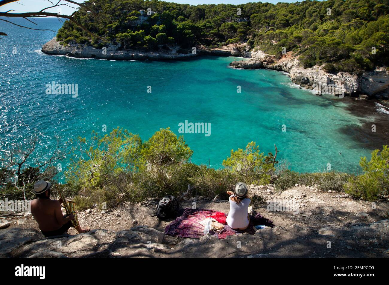 a man plays his saxaphone looking down on cala mitjanetta from the cliffs above cala mitjana menorca spain Stock Photo
