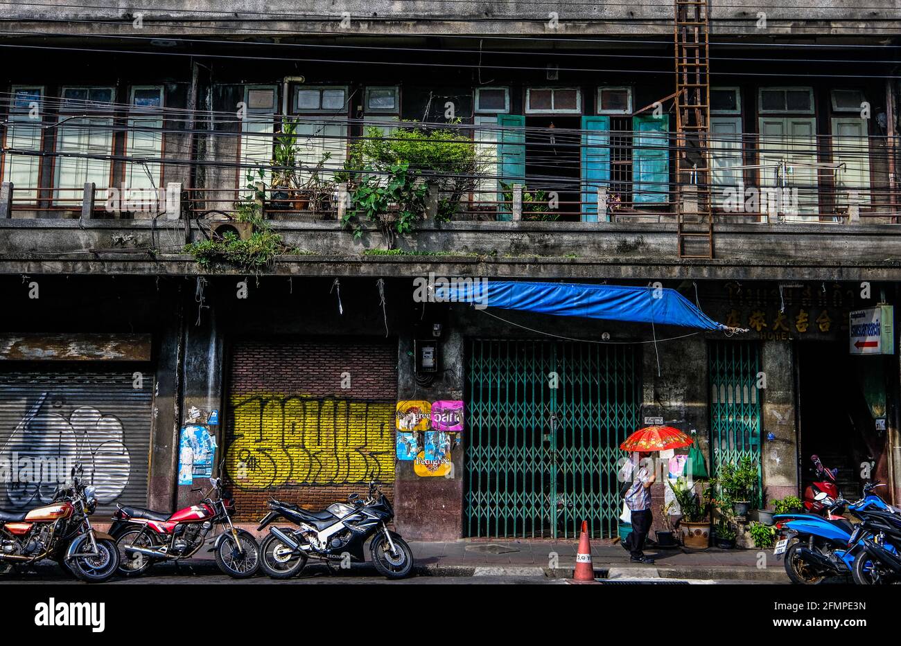 A man with a red umbrella walks past an old, graffiti-clad, building in the Chinatown area of Bangkok, Thailand Stock Photo