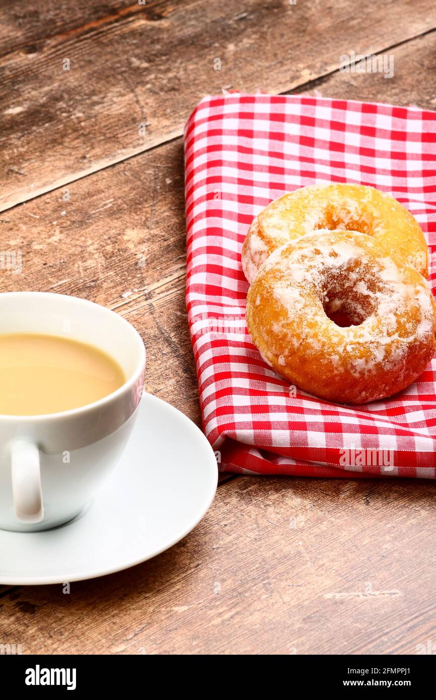 Two tasty ring doughnuts sat on a red and white checked cloth on a wooden table top with a cup of tea Stock Photo