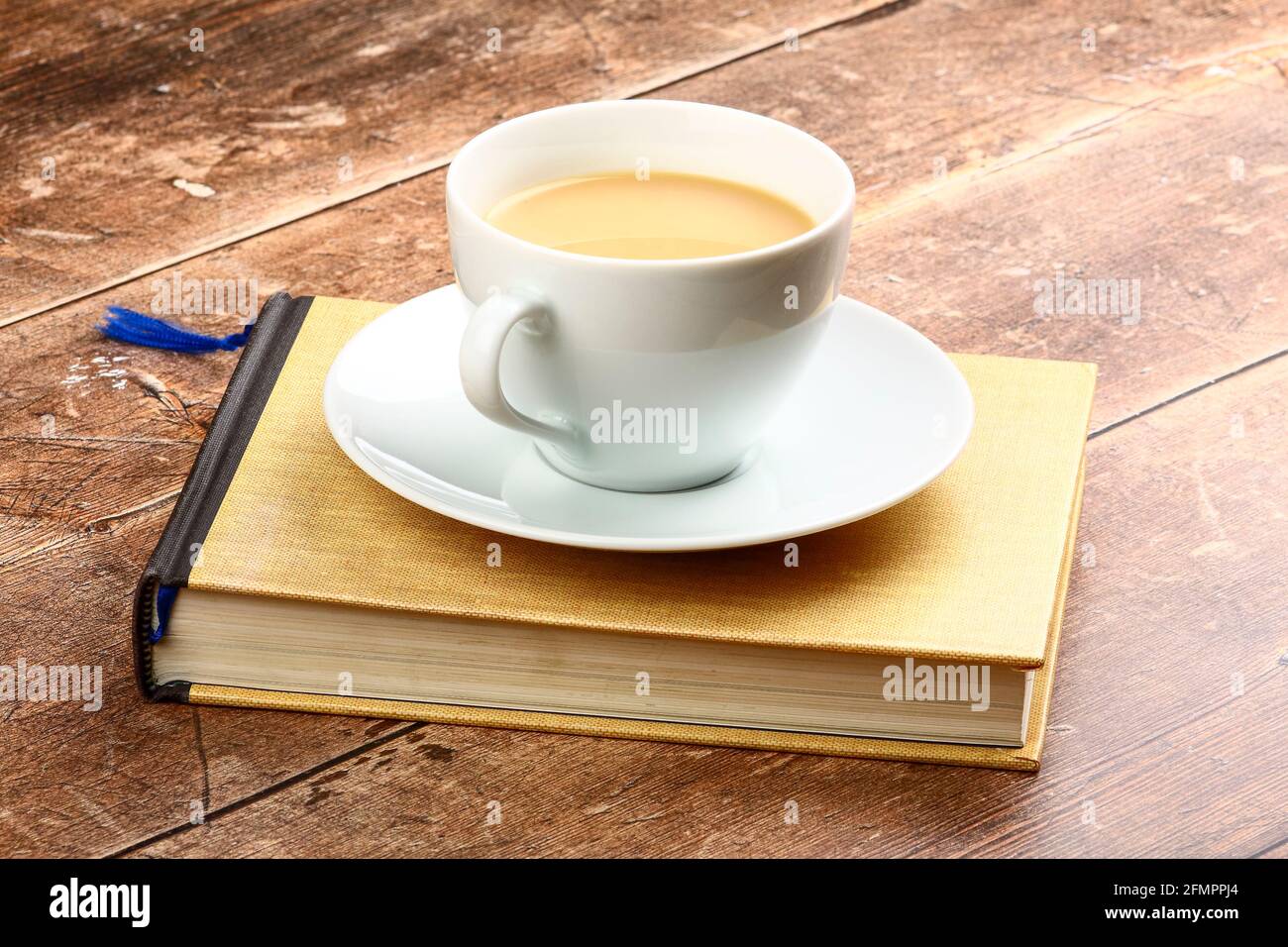 White cup and saucer filled with tea sat on a reading book on a rustic wooden table top Stock Photo