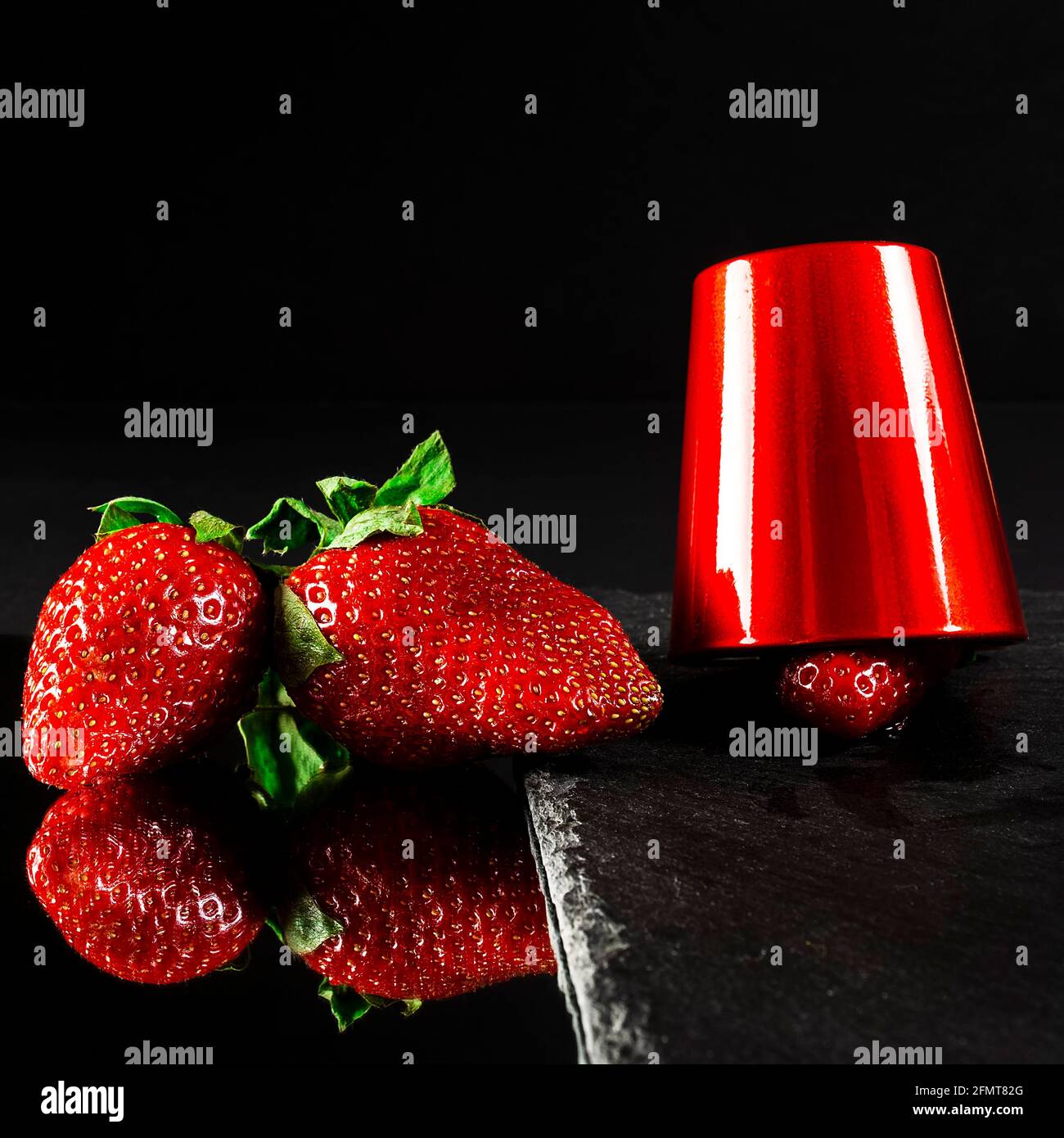 Two fresh red strawberries reflected on a mirror and a red strawberry under a red cup on a slate base.The photograph is a still life taken with artifi Stock Photo