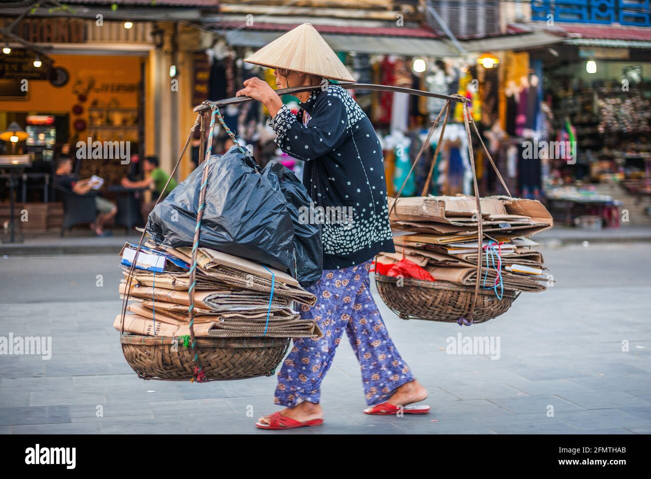 Elderly Vietnamese lady wearing conical hat carries twin baskets on shoulder pole containing folded cardboard boxes, Hoi An, Vietnam Stock Photo