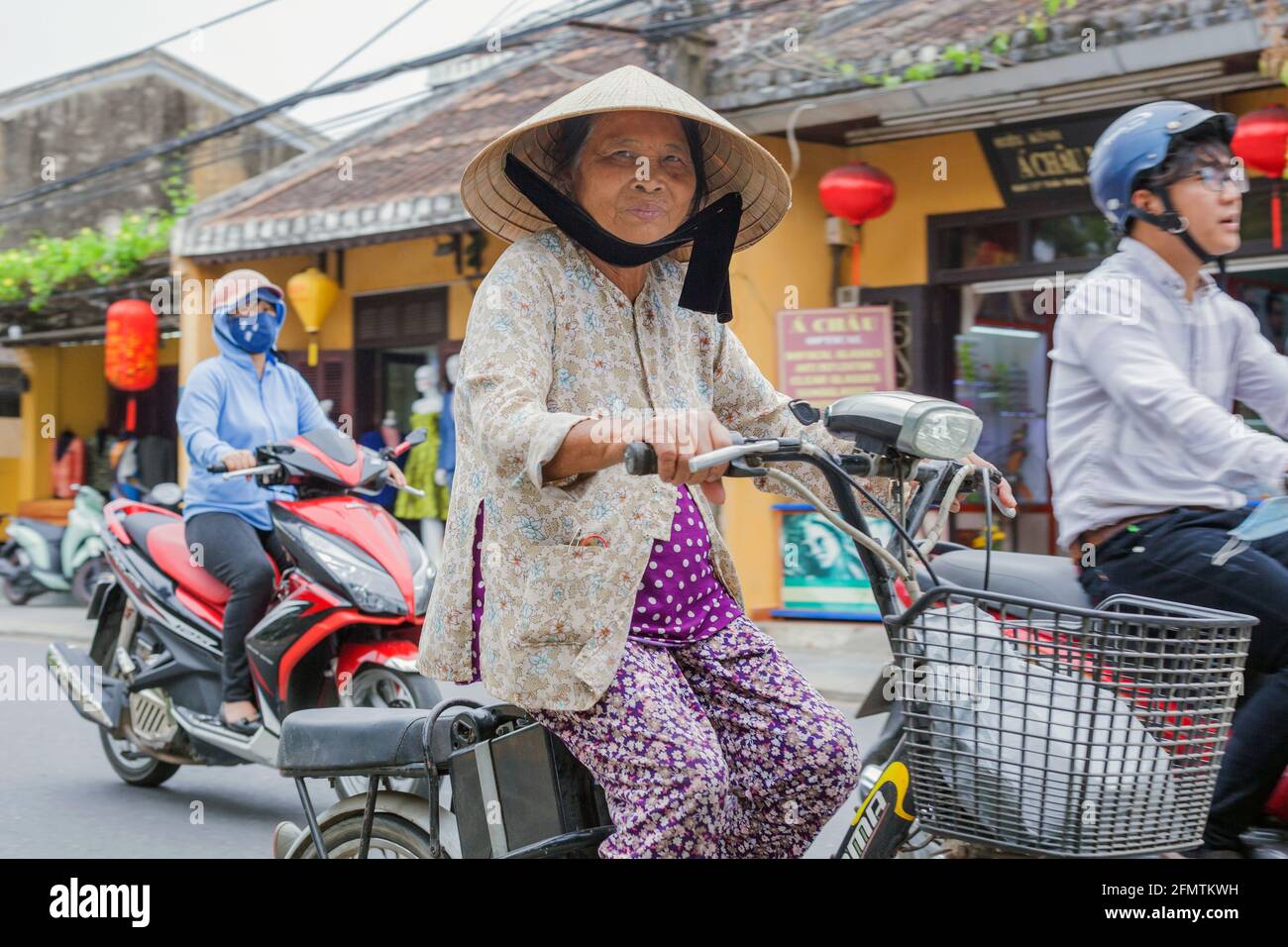 Elderly Vietnamese lady wearing conical hat rides bicycle through the old town, Hoi An, Vietnam Stock Photo