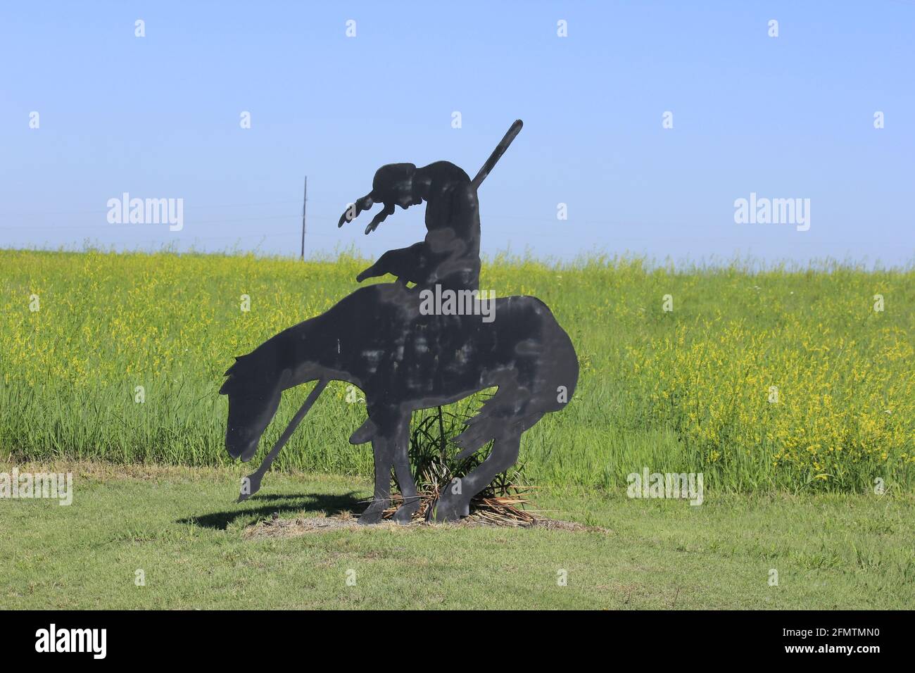 A Metal Indian cut out that's black with a farm field in the background with weeds with yellow flowers in Kansas. Stock Photo