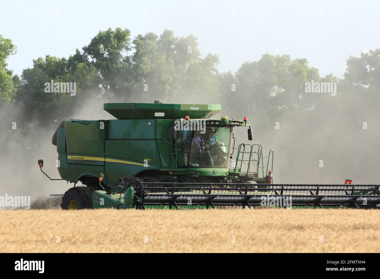 John Deere Combine cutting wheat with wheat dust in the air  in a farm field out in the country in Kansas. Stock Photo