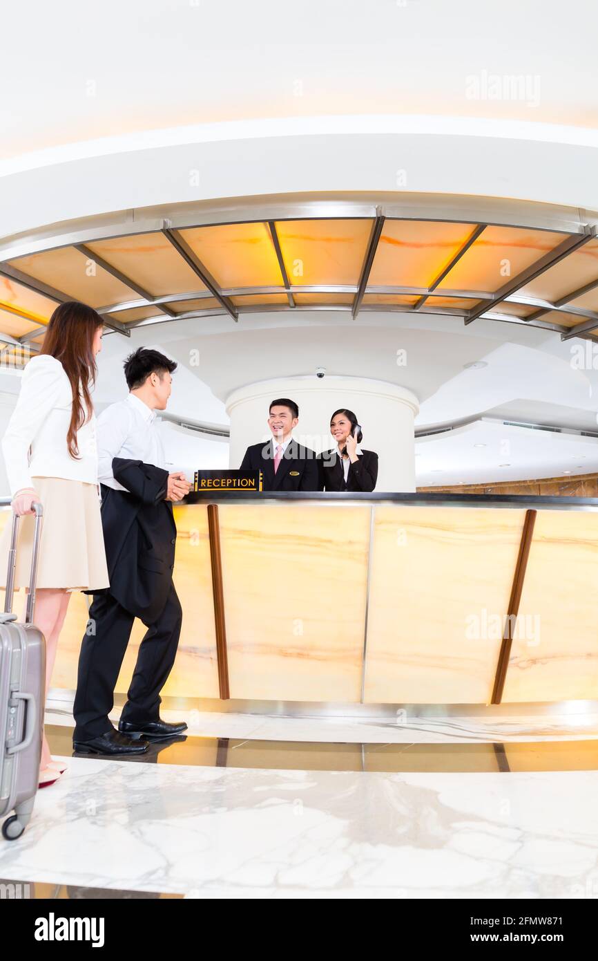 Asian Chinese woman and man arriving at front desk or reception of luxury hotel in business clothes with trolley Stock Photo