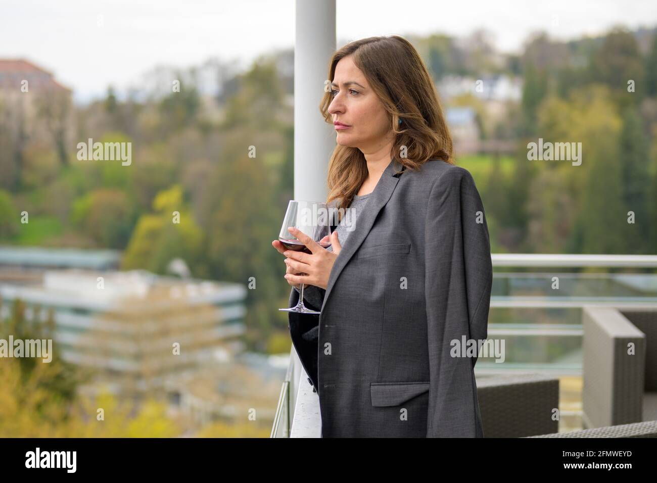 Serious withdrawn middle-aged woman looking aside as she stands on a balcony with a glass of wine and her husbands suit jacket over her shoulders Stock Photo
