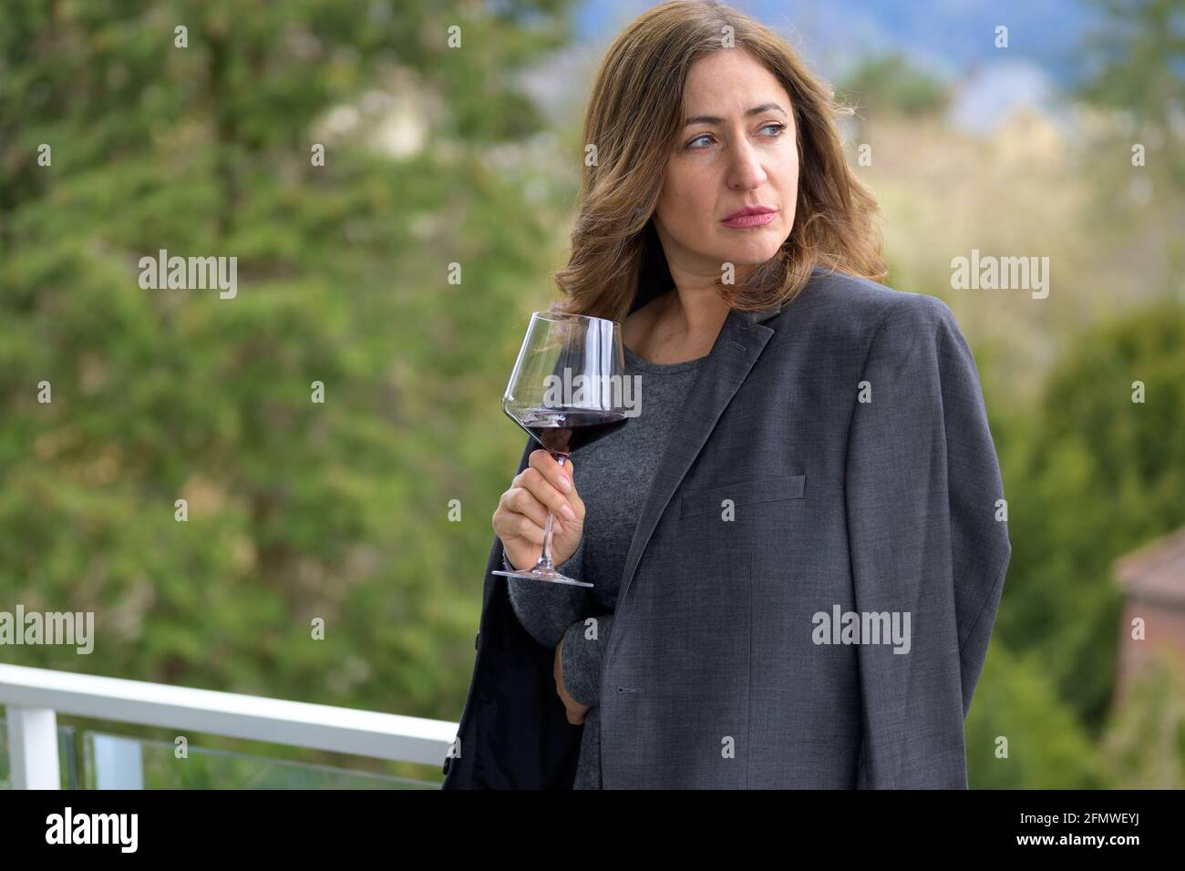 Serious withdrawn middle-aged woman looking aside as she stands on a balcony with a glass of wine and her husbands suit jacket over her shoulders Stock Photo