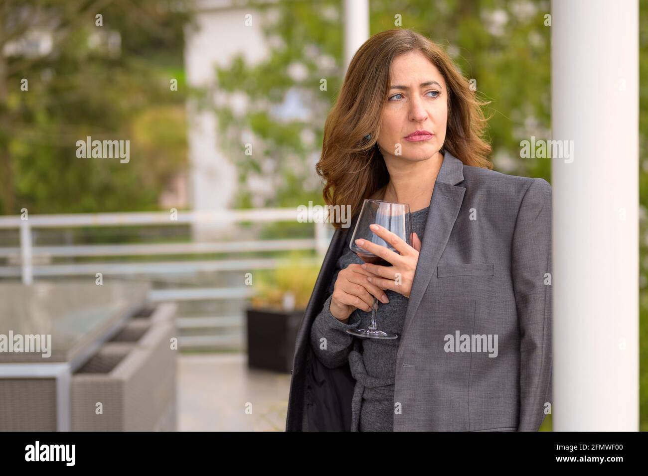 Woman relaxing on a balcony with a glass of wine standing leaning against therailing with a jacket draped over her shoulders looking pensively aside Stock Photo