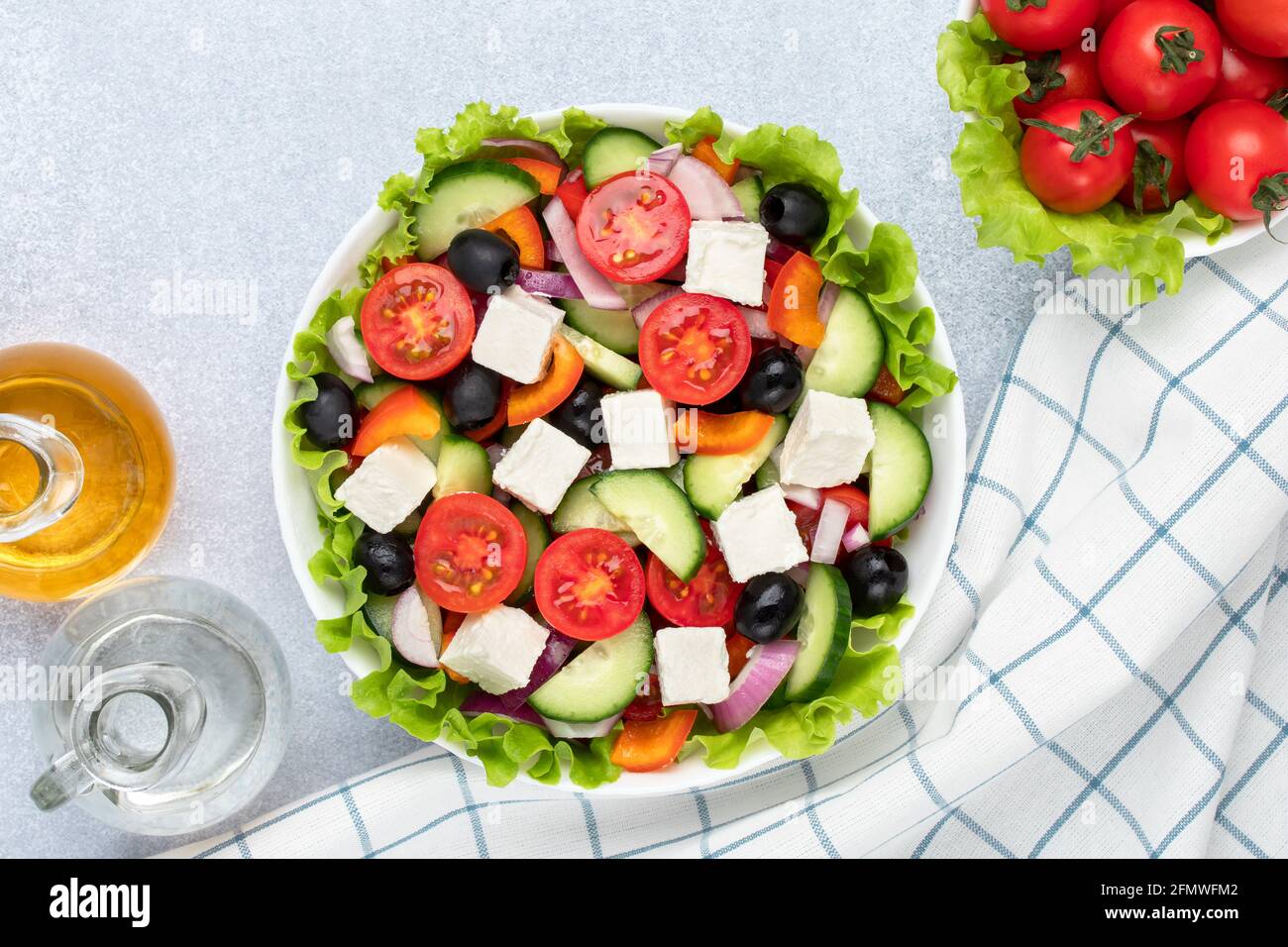 Greek salad with cherry tomatoes, cucumbers, feta cheese, olives and seasoned with oil. White checkered napkin on a gray table. Top view. Flat lay com Stock Photo