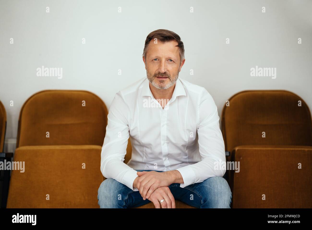 Intense middle aged man staring at the camera as he sits forwards on a row of seats against a white wall with a serious determined expression Stock Photo
