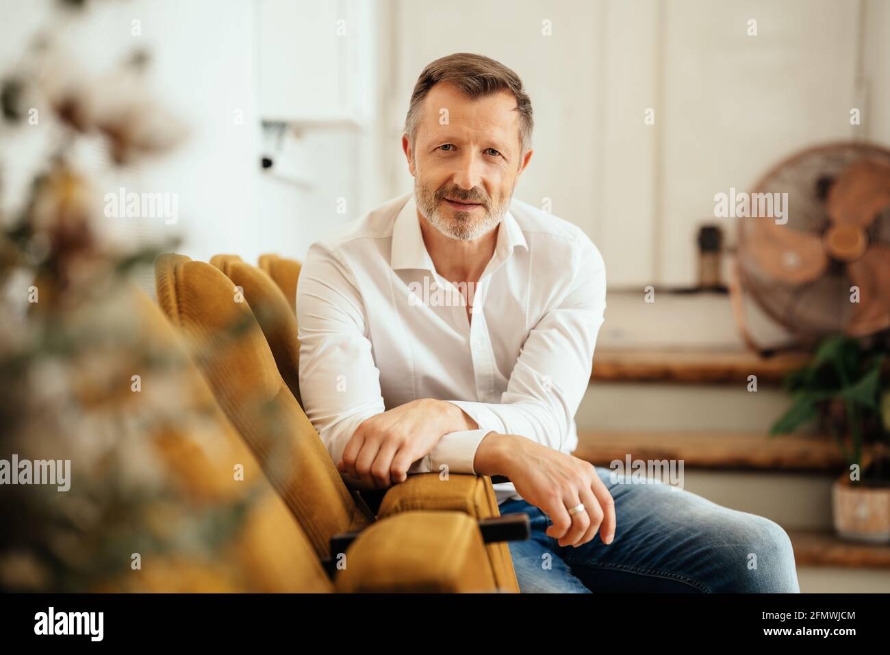 Relaxed casual trendy middle-aged man staring at the camera as he turns to lean on a row of seats against a wall Stock Photo