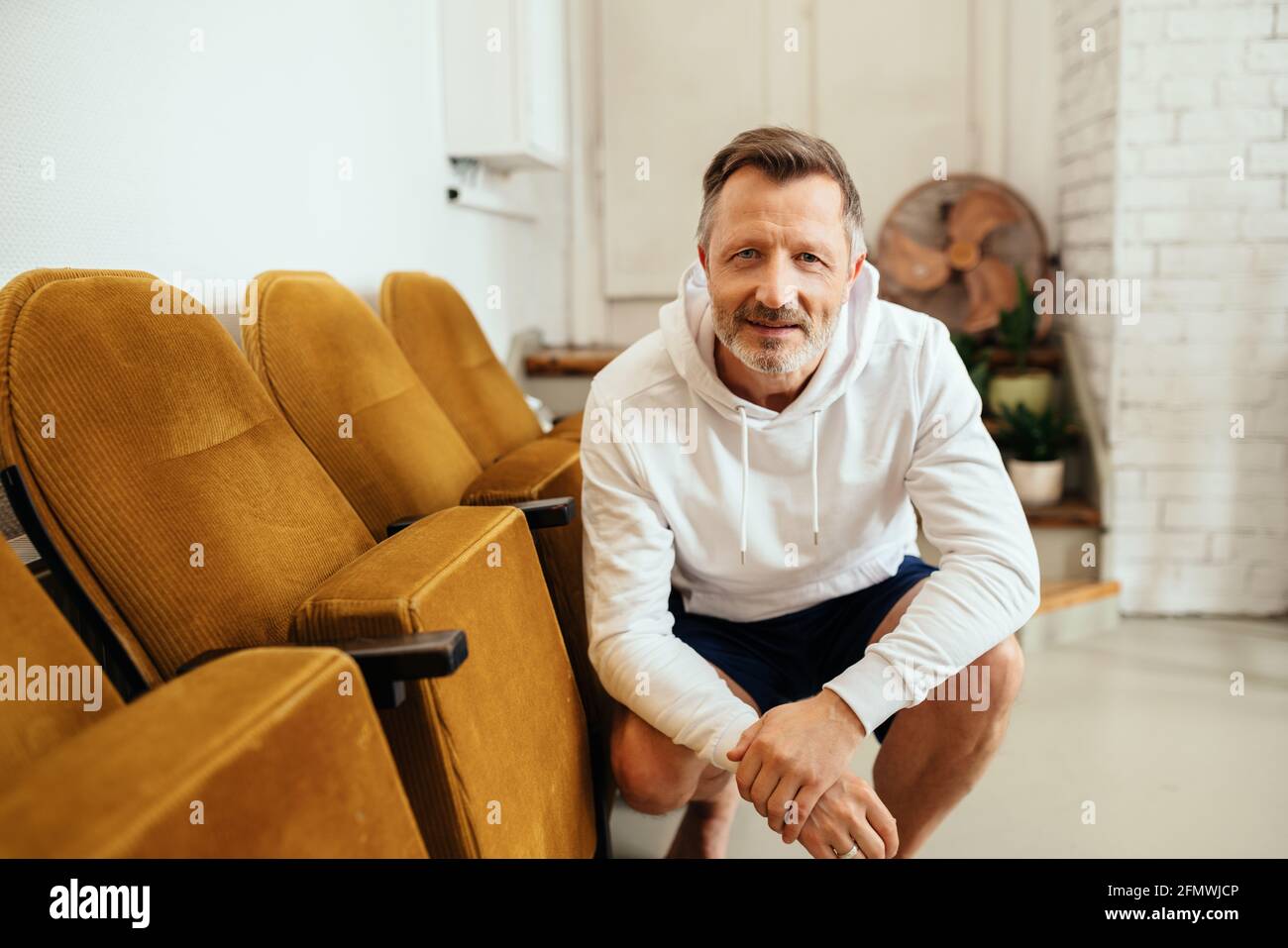Middle-aged man in shorts and a white hoodie kneeling staring at the camera with an expectant attentive expression as though listening Stock Photo