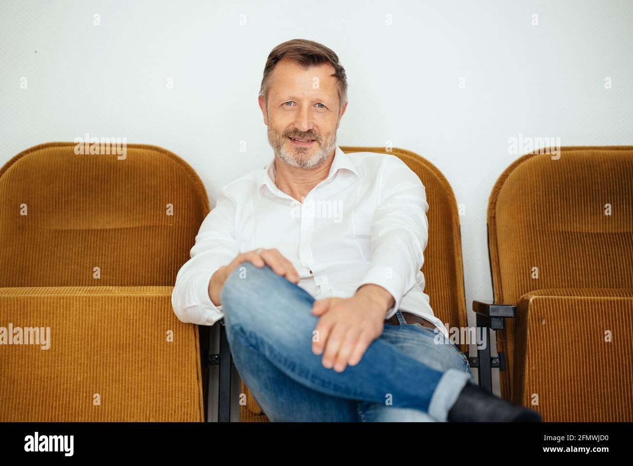 Relaxed confident middle-aged man smiling quietly t the camera as he sits in jeans and white shirt on a row of chairs against a wall with copyspace Stock Photo